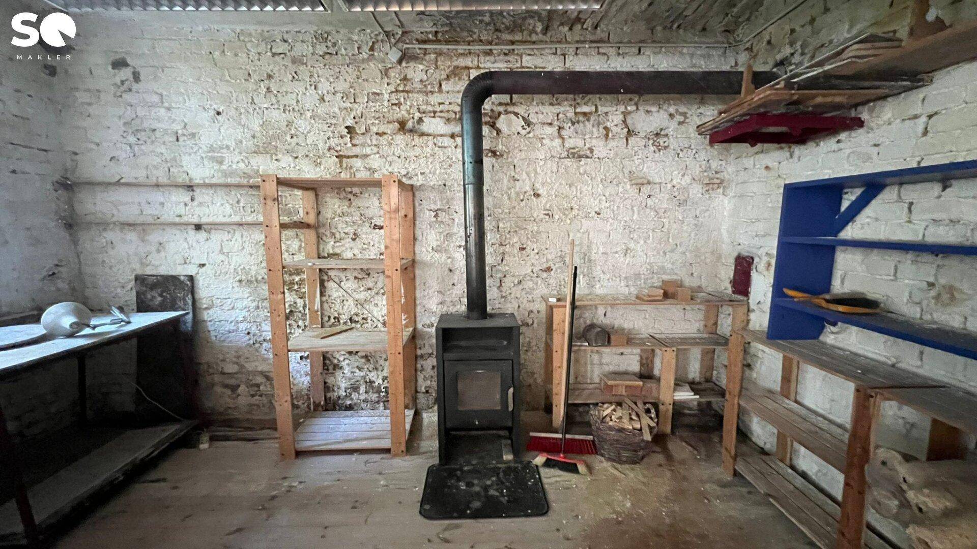 Rustikaler Kellerraum mit freistehendem Holzofen, unverputzten Ziegelwänden und Holzregalen, ideal für Lagerzwecke.