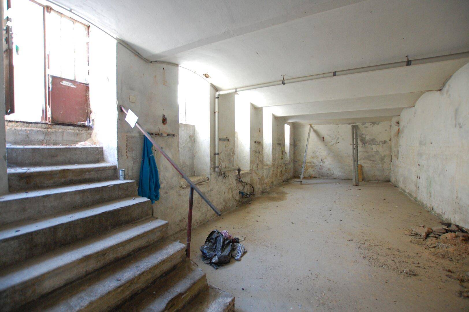 Unfertiger Kellerraum mit Betonboden, kleinen Fenstern und einer Betontreppe, die nach oben führt.