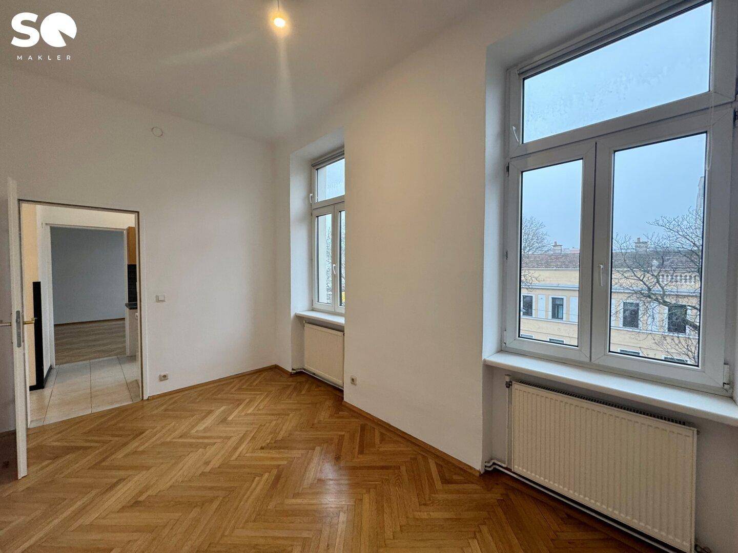 Helles Wohnzimmer mit Fischgrätparkett und hohen Fenstern, bietet Zugang zu einem weiteren Raum.