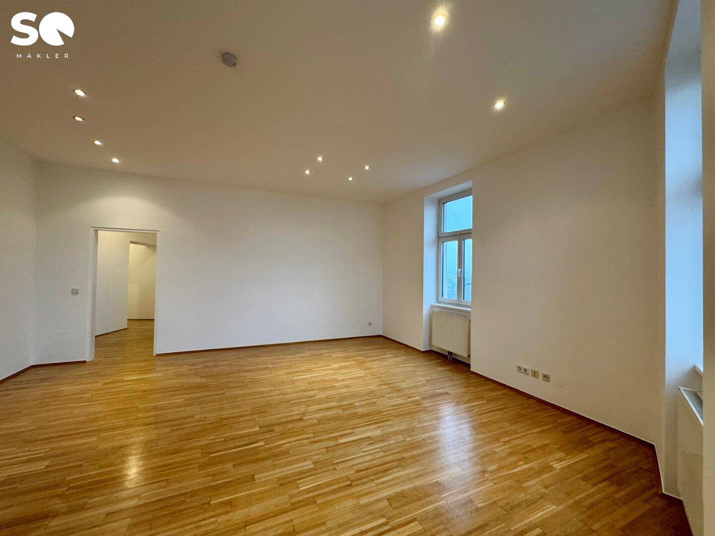 Geräumiges, leeres Wohnzimmer mit Parkettboden, Einbaustrahlern und Blick auf den Flur.