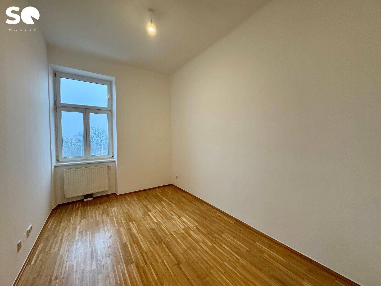 Helles Schlafzimmer mit Parkettboden, einem Fenster und weiß gestrichenen Wänden.