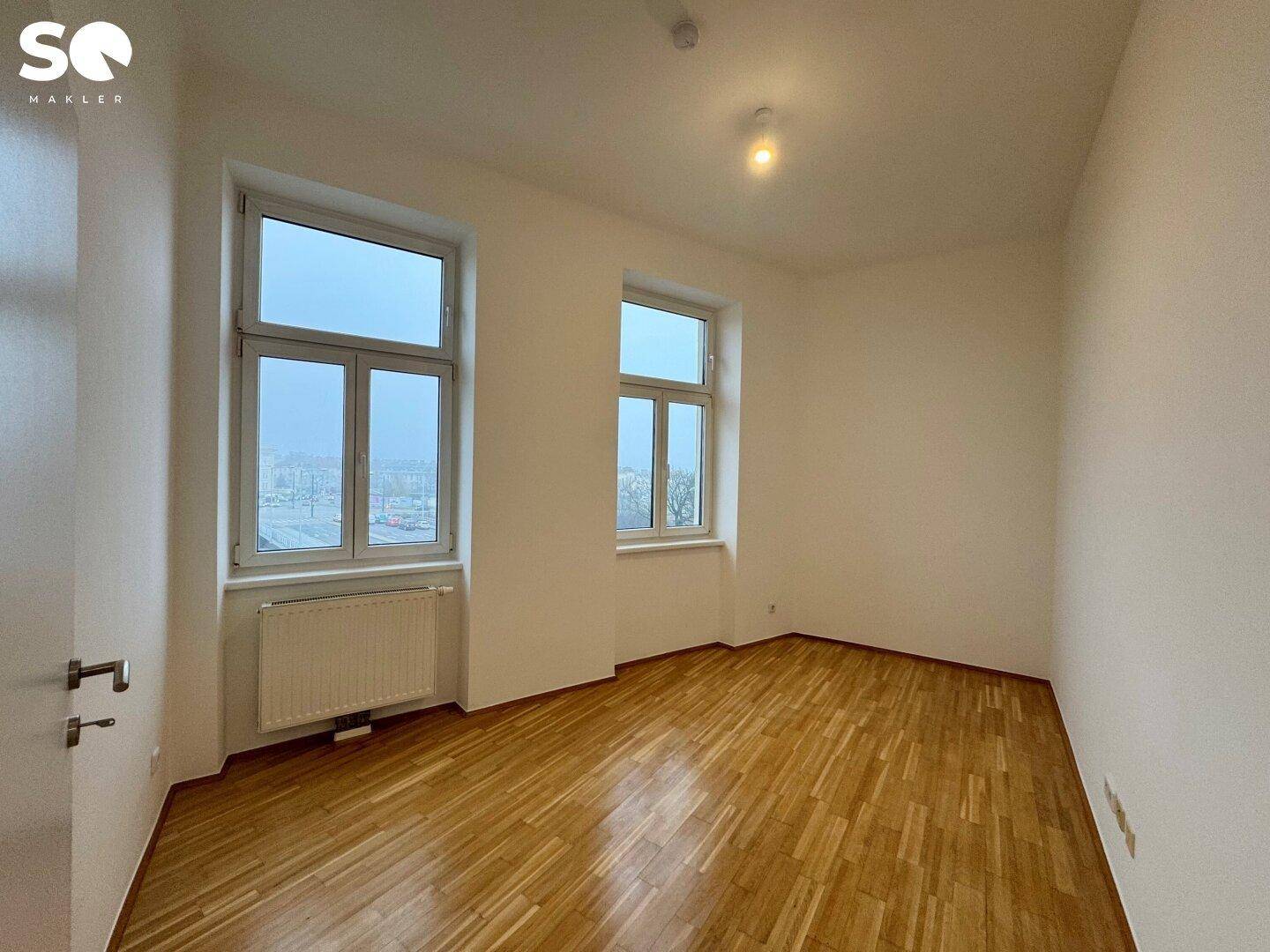 Leeres Schlafzimmer mit zwei Fenstern, Parkettboden und Heizkörpern unter den Fenstern.