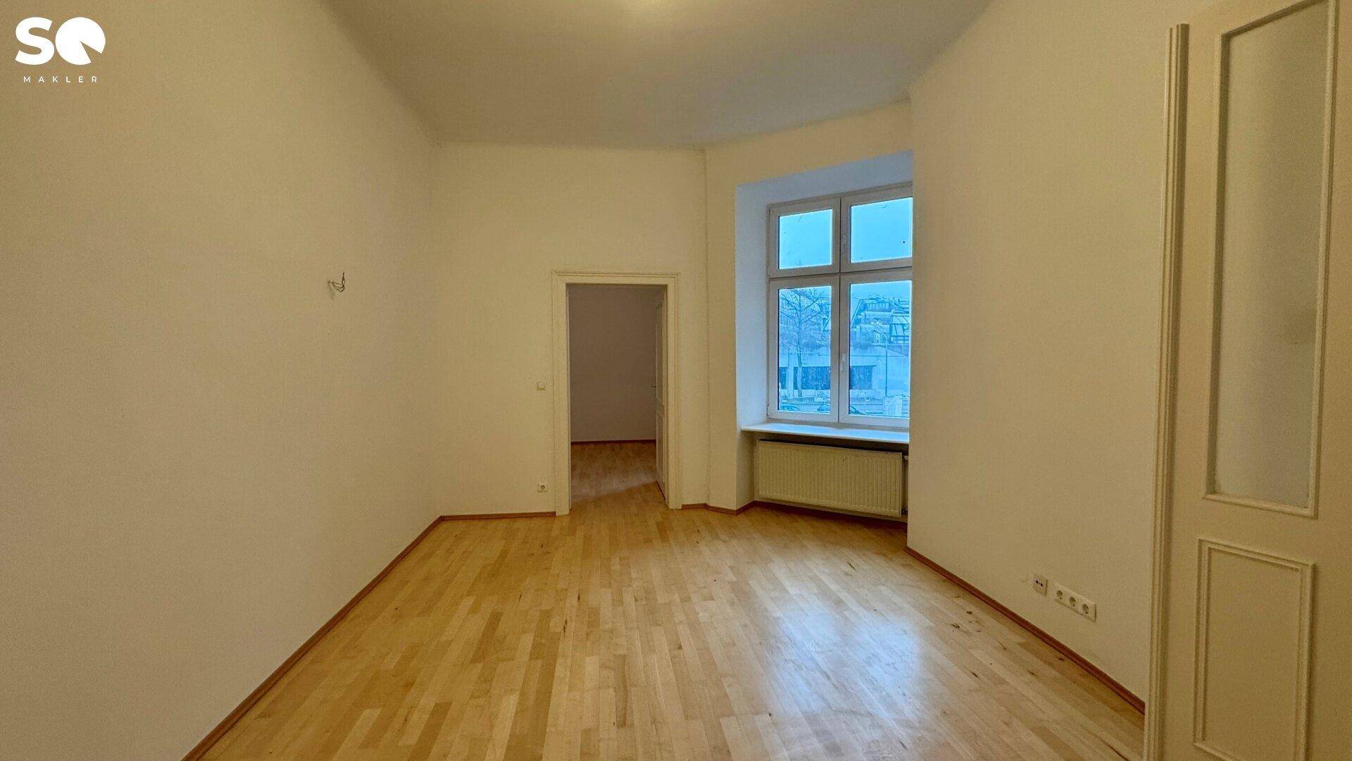 Großes, leeres Zimmer mit Parkettboden und einem Fenster, das den Raum mit Tageslicht füllt.