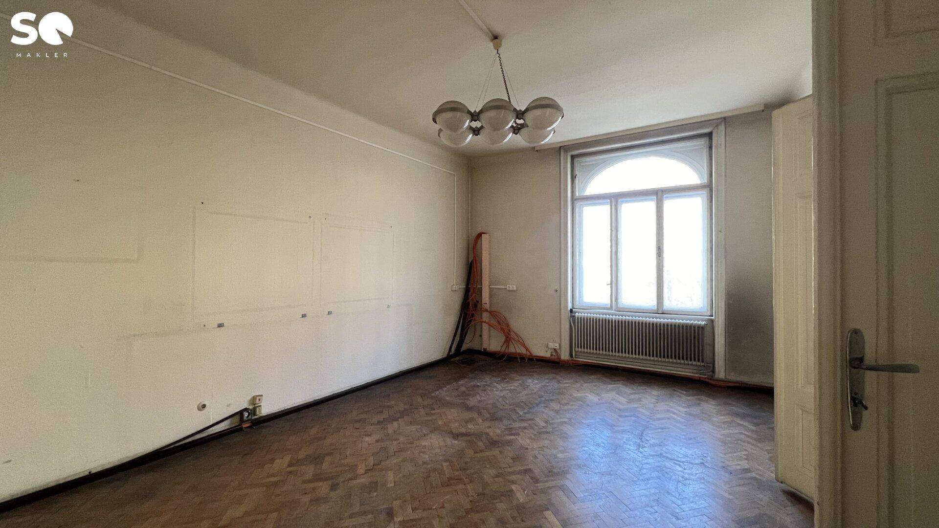 Geräumiges Zimmer mit klassischem Fischgrätparkett, einem großen Bogenfenster und einer Deckenleuchte.
