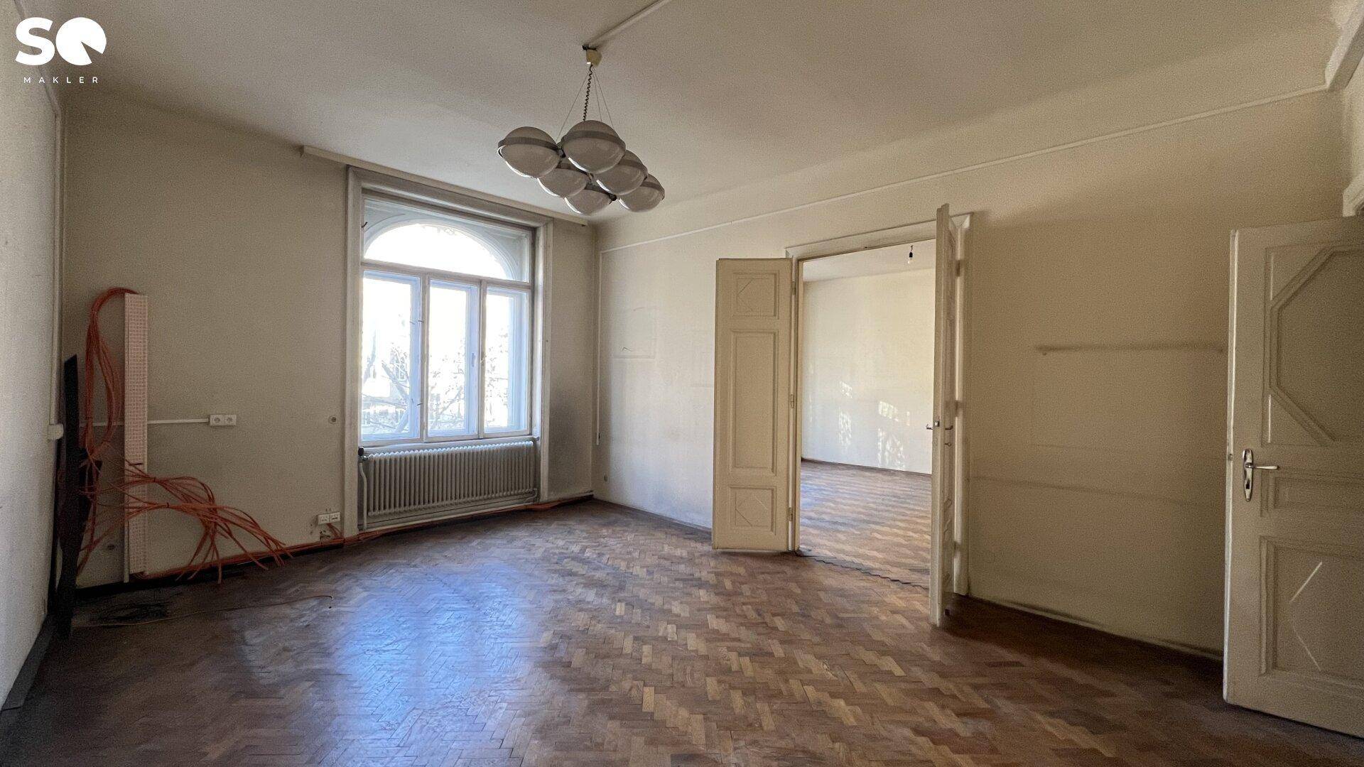Geräumiges Zimmer mit Fischgrätparkett, hohen Decken und einem großen Bogenfenster, bereit zur Renovierung.