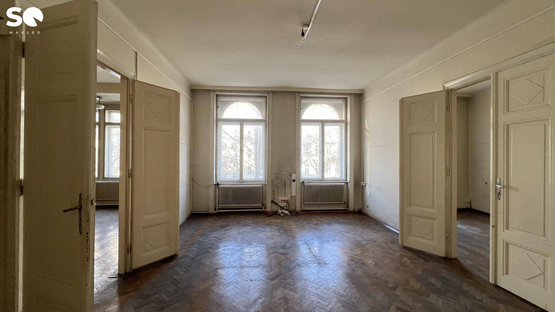 Großes Zimmer mit zwei hohen Bogenfenstern, Fischgrätparkett und Zugang zu weiteren Räumen.