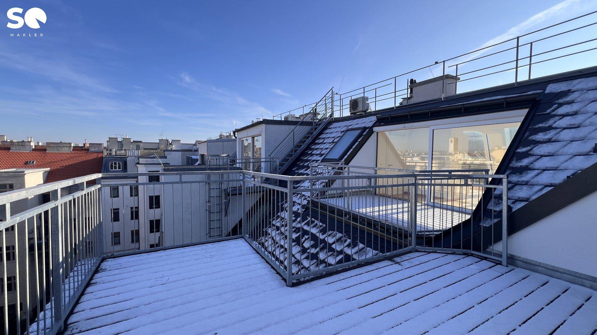 Große Dachterrasse mit Schnee bedeckt, umgeben von Stadtgebäuden unter blauem Himmel.