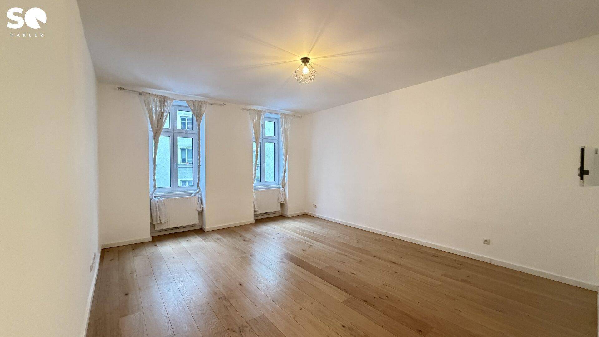 Geräumiges Wohnzimmer mit hellem Parkettboden und zwei großen Fenstern für viel Tageslicht.