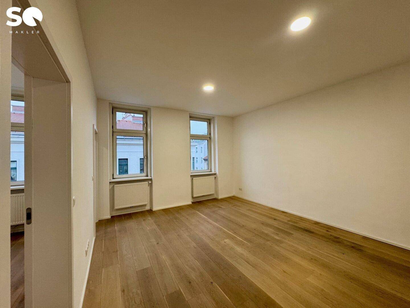 Helles, leeres Zimmer mit Holzboden und zwei Fenstern, das viel Tageslicht hereinlässt.