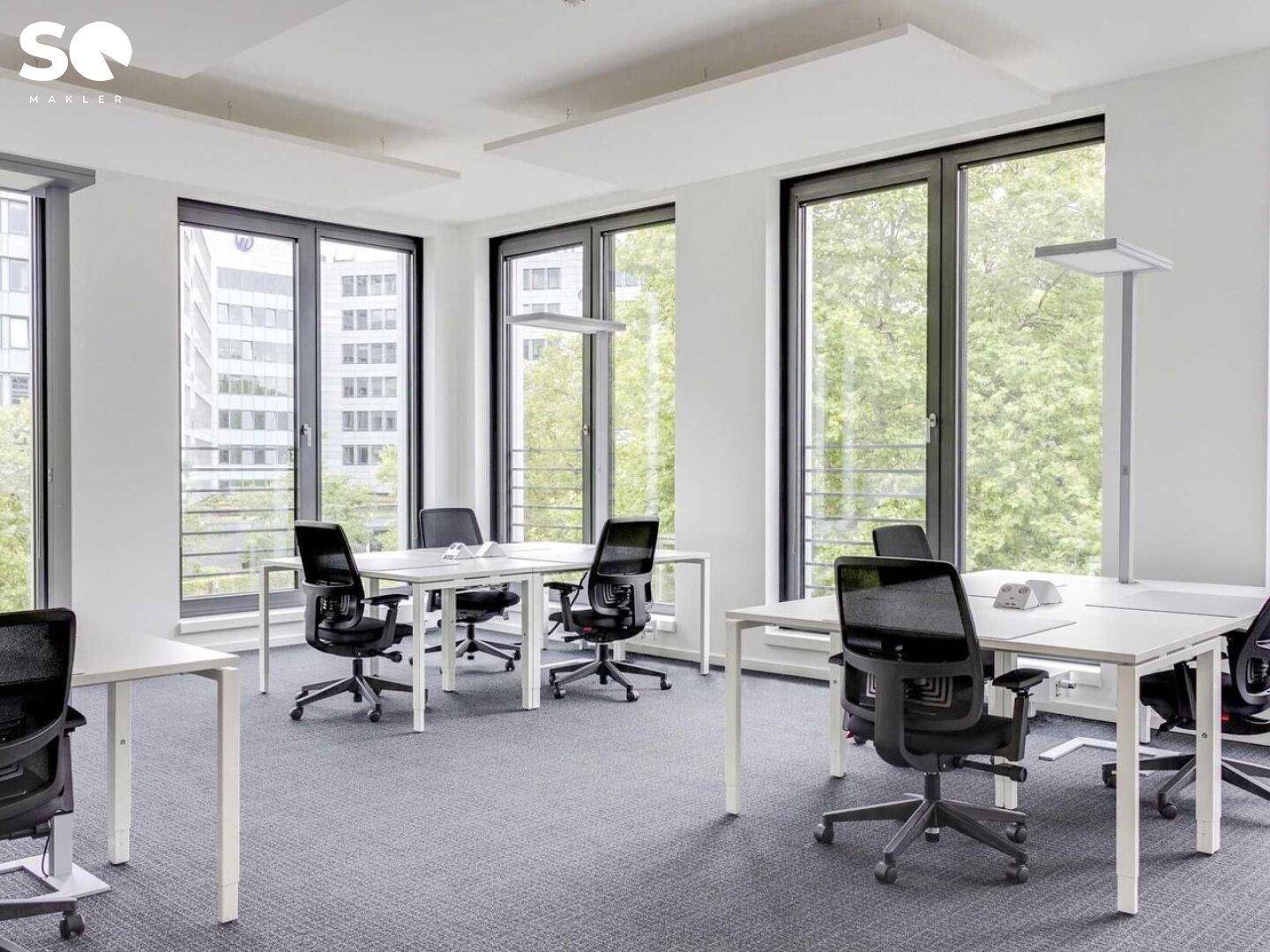 Helles Büro mit mehreren Schreibtischen, ergonomischen Stühlen und großen Fenstern mit Blick ins Grüne.
