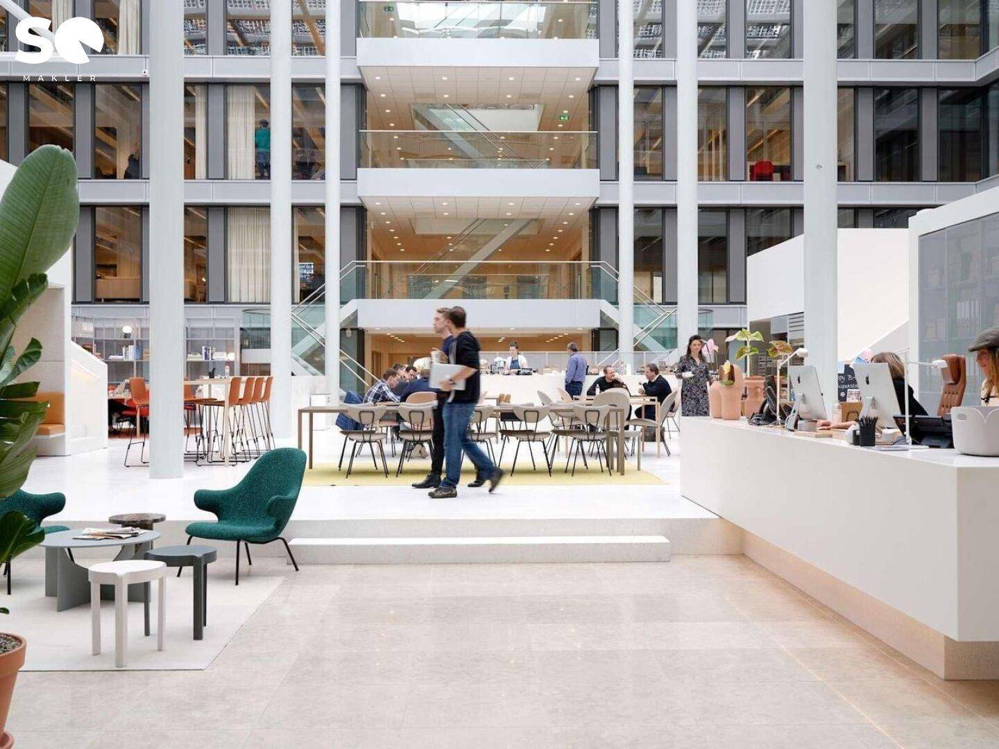 Großzügiges, mehrstöckiges Büroatrium mit Empfangsbereich, Lounge-Zonen und vielen Menschen.