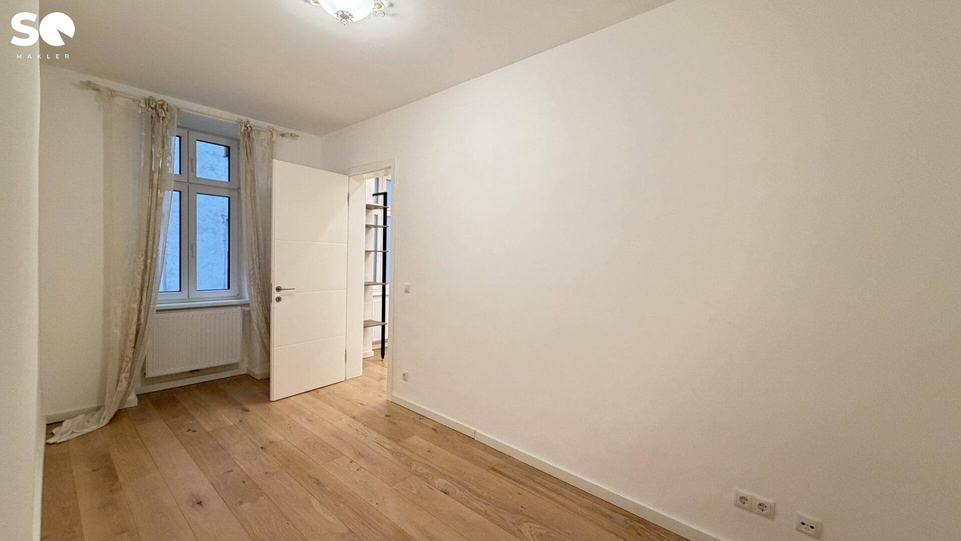 Helles Schlafzimmer mit Parkettboden und Fenster, das einen Blick in den angrenzenden Raum bietet.