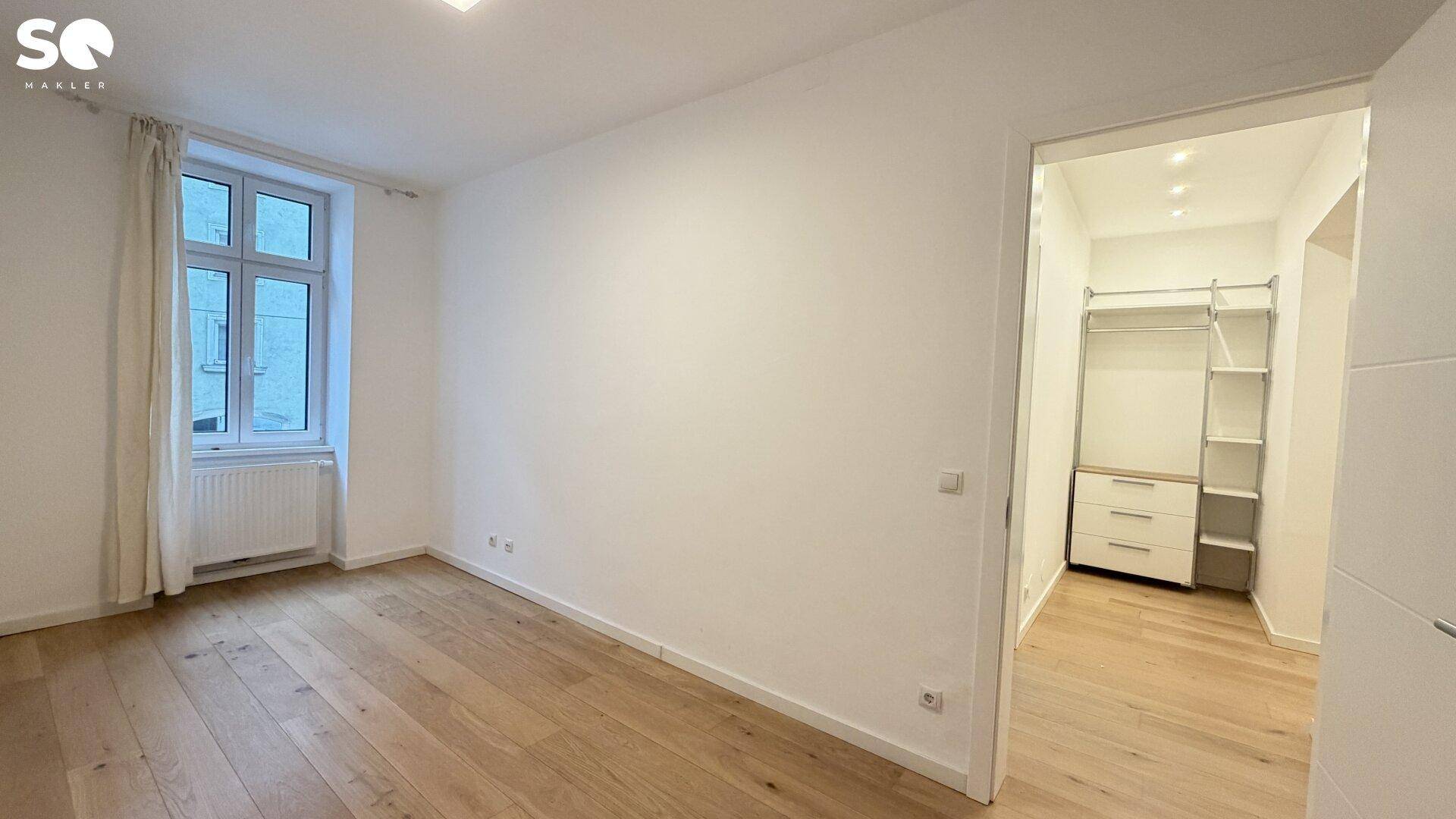 Helles Zimmer mit Echtholzboden, großem Fenster und direktem Zugang zu einem begehbaren Kleiderschrank.