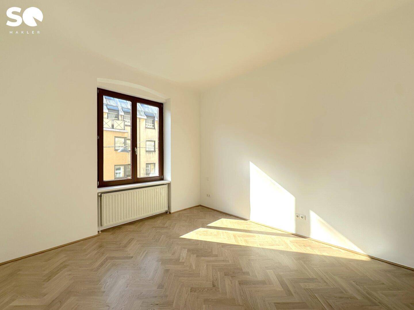 Freundliches Zimmer mit hellem Fischgrätparkett und einem großen Fenster, das viel Tageslicht hereinlässt.