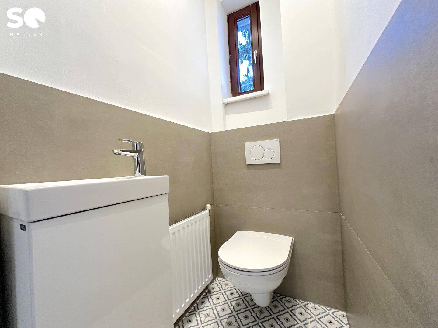 Gäste-WC mit modernem Wand-WC, kleinem Waschbecken und einem Fenster für natürliche Belüftung.