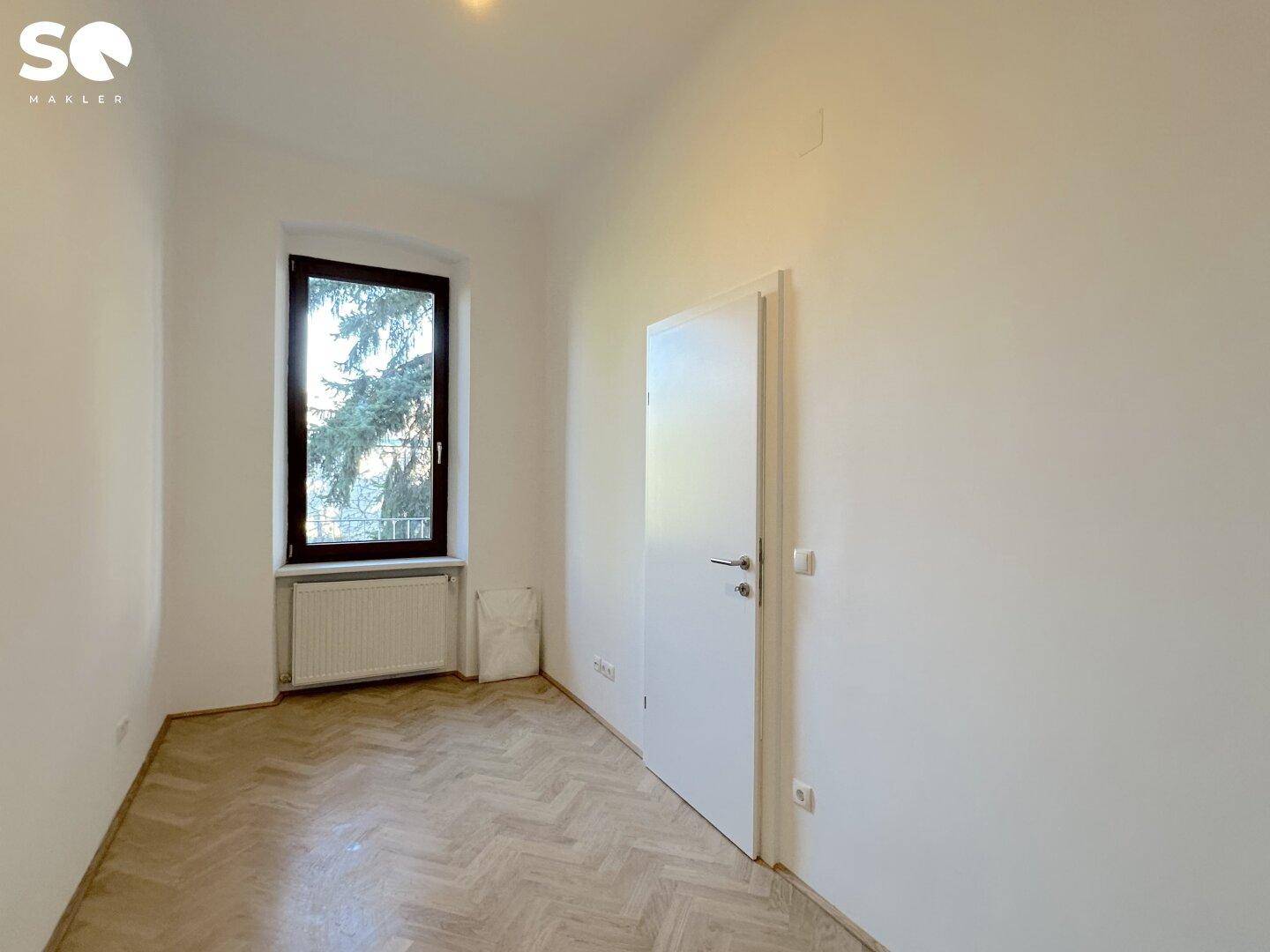 Ruhiges Schlafzimmer mit hellem Fischgrätparkett, weißen Wänden und einem Fenster mit Blick ins Grüne.