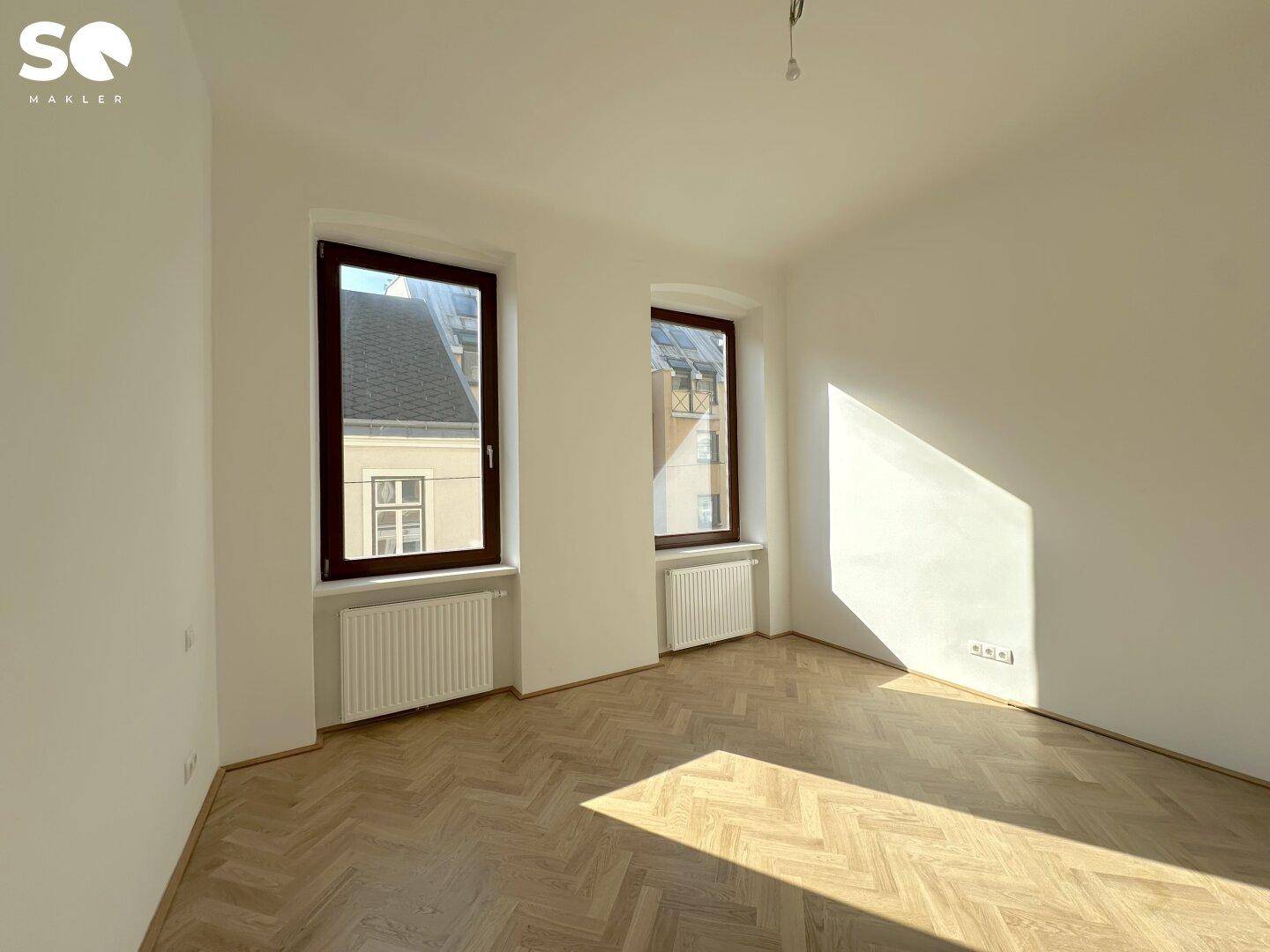 Helles Wohnzimmer mit elegantem Fischgrätparkett und weißen Wänden, durchflutet von natürlichem Sonnenlicht.