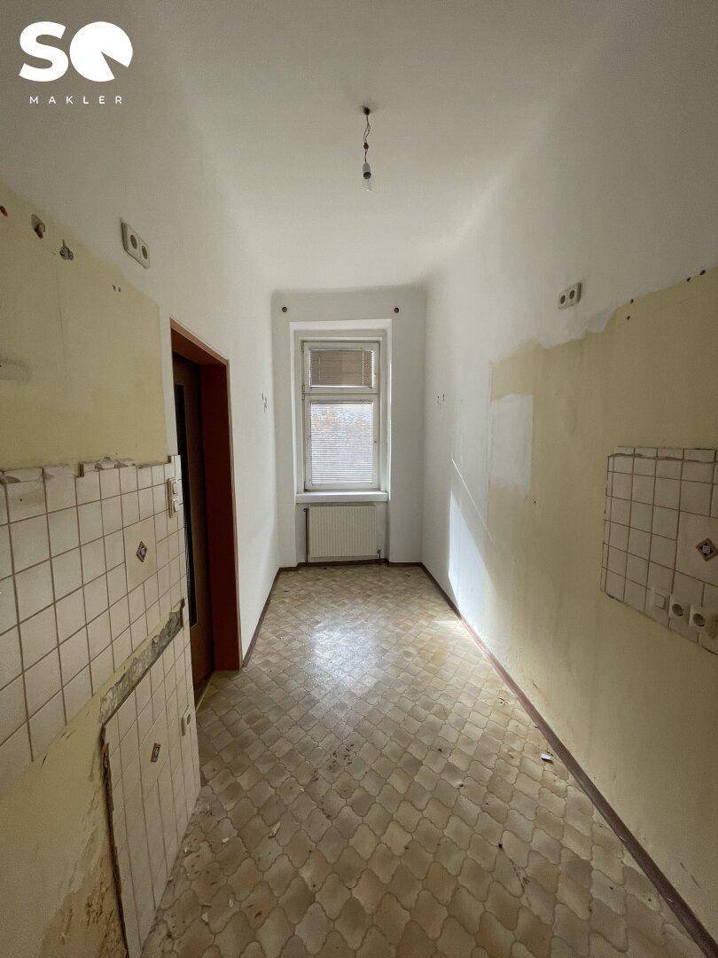 Langer Flur mit alten Fliesen und teilweise abgenutzten Wänden, der zu einem Fenster führt.