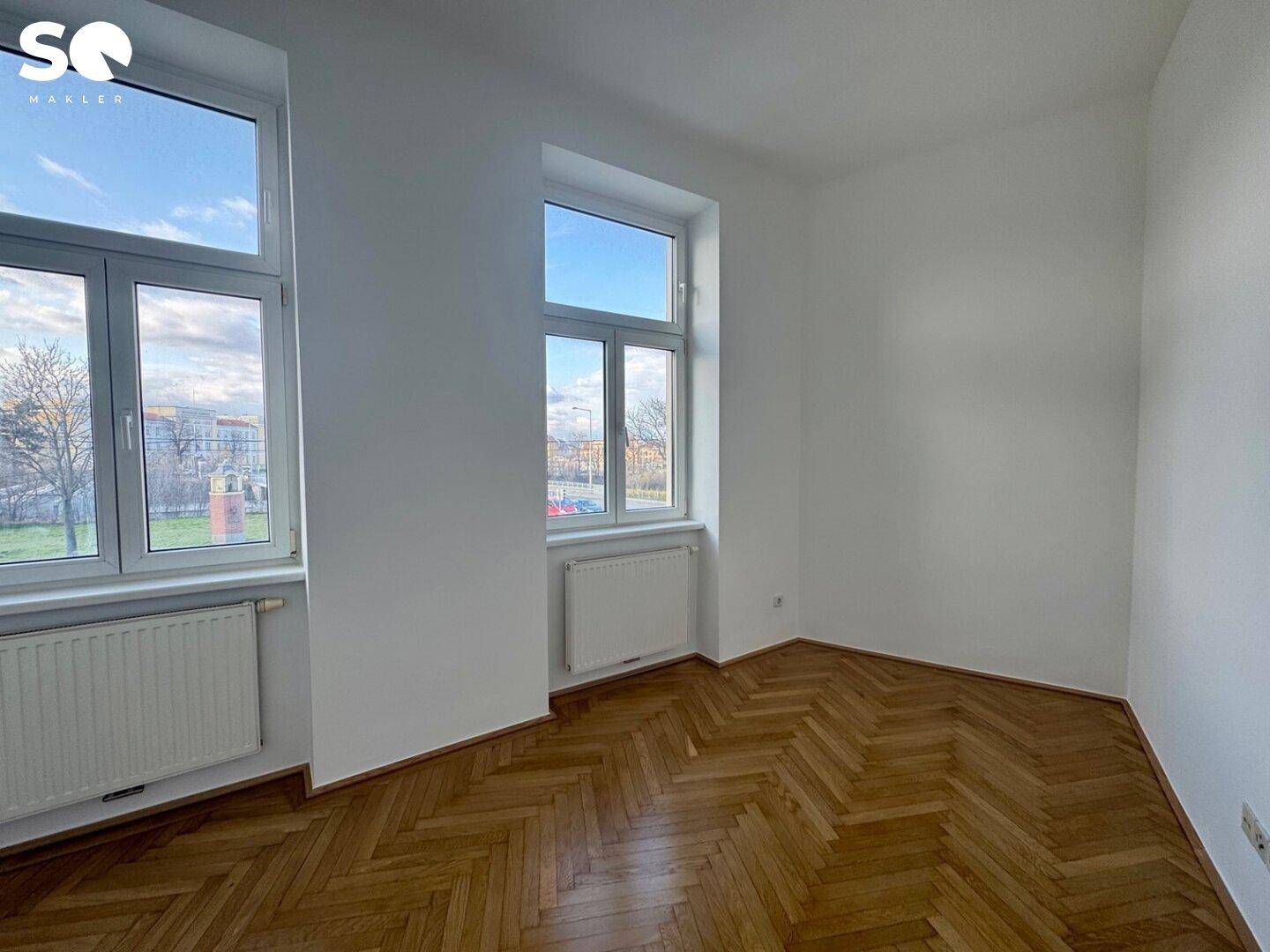 Helles Zimmer mit Fischgrätparkett und zwei Fenstern, die viel Licht hereinlassen.