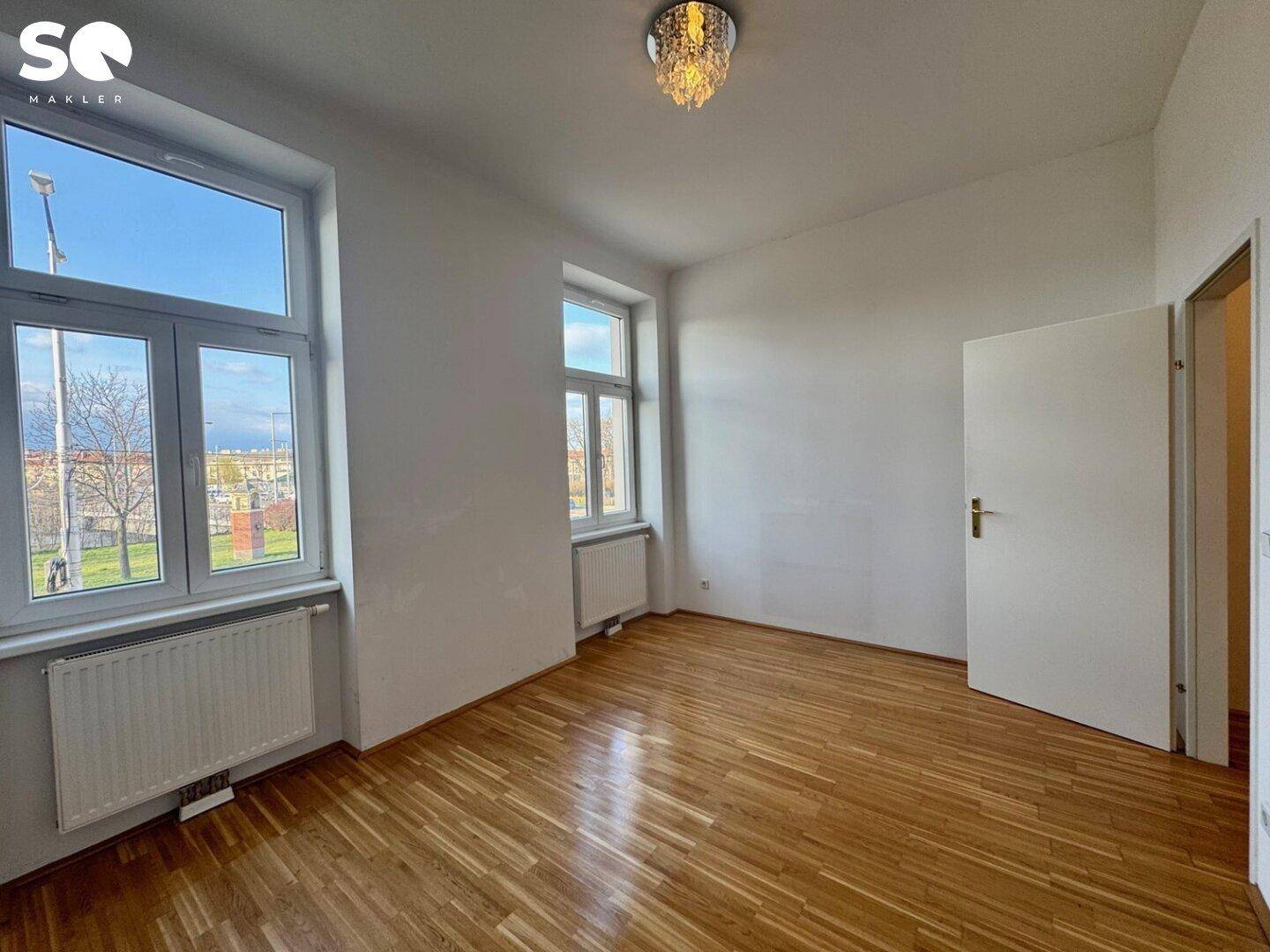 Geräumiges, helles Zimmer mit Holzboden und Blick nach draußen, ideal als Wohn- oder Schlafzimmer nutzbar.