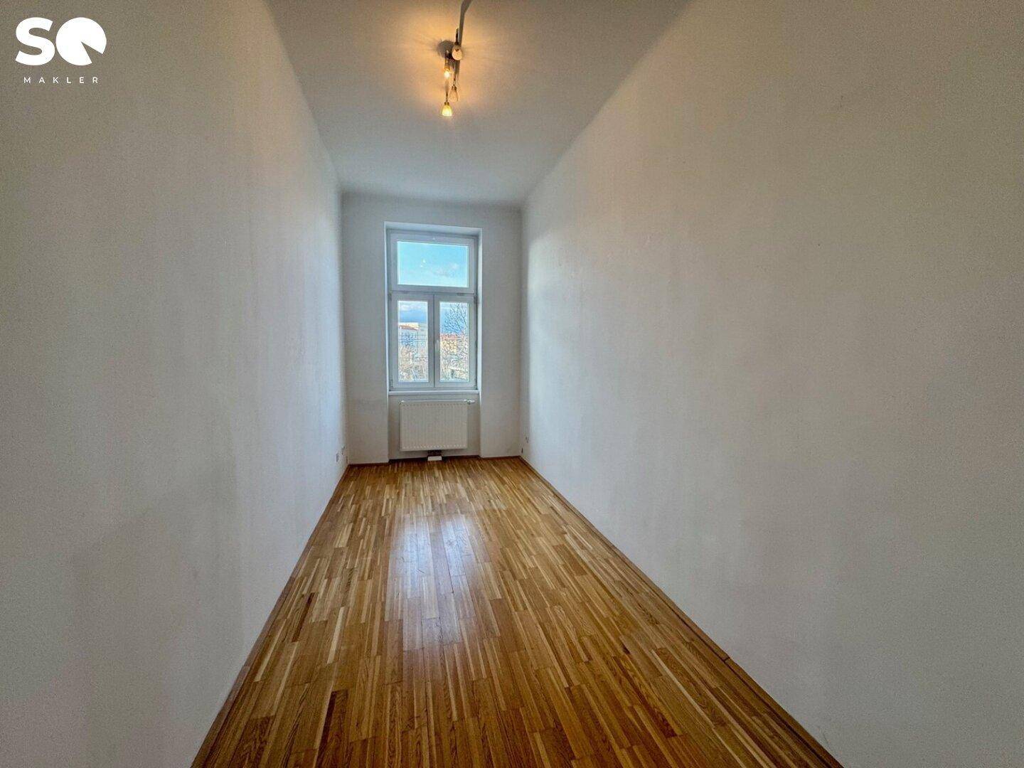 Langes, schmales Zimmer mit Holzboden und einem Fenster, das den Raum mit natürlichem Tageslicht versorgt.
