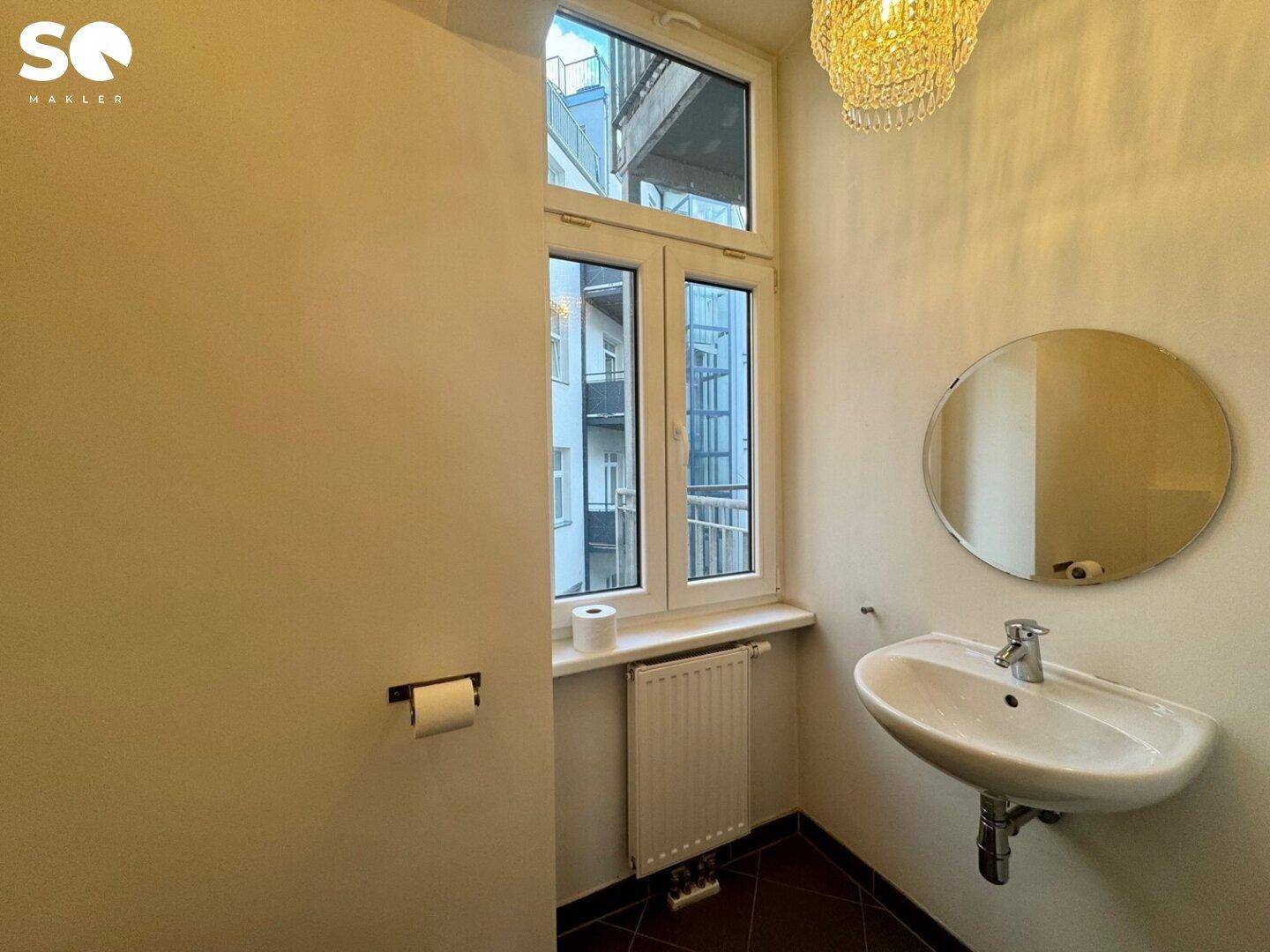 Kleines Gäste-WC mit Waschbecken, Fenster und einem runden Spiegel, hell und zweckmäßig eingerichtet.