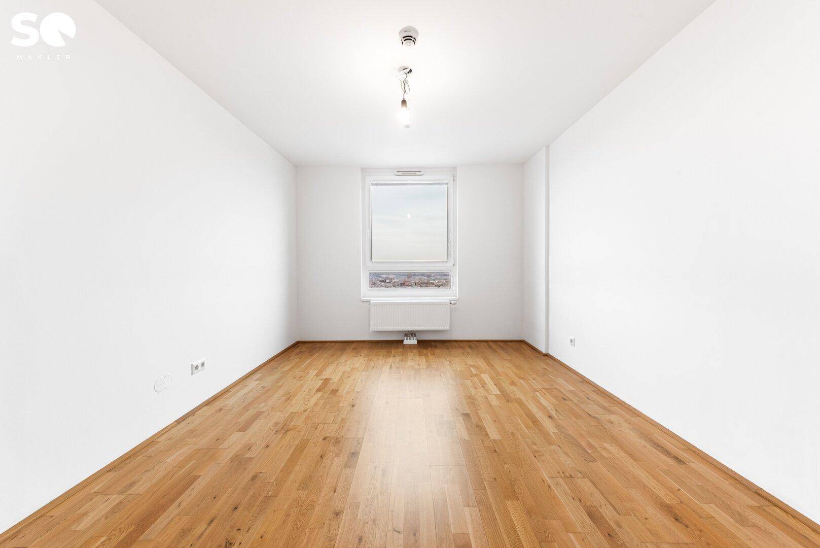 Leeres Schlafzimmer mit Parkettboden, weißen Wänden und Fenster mit Stadtblick.