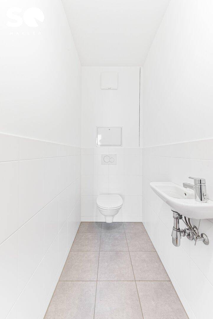 Separates WC mit Wand-WC, kleinem Waschbecken und hellen Fliesen an Wänden und Boden.