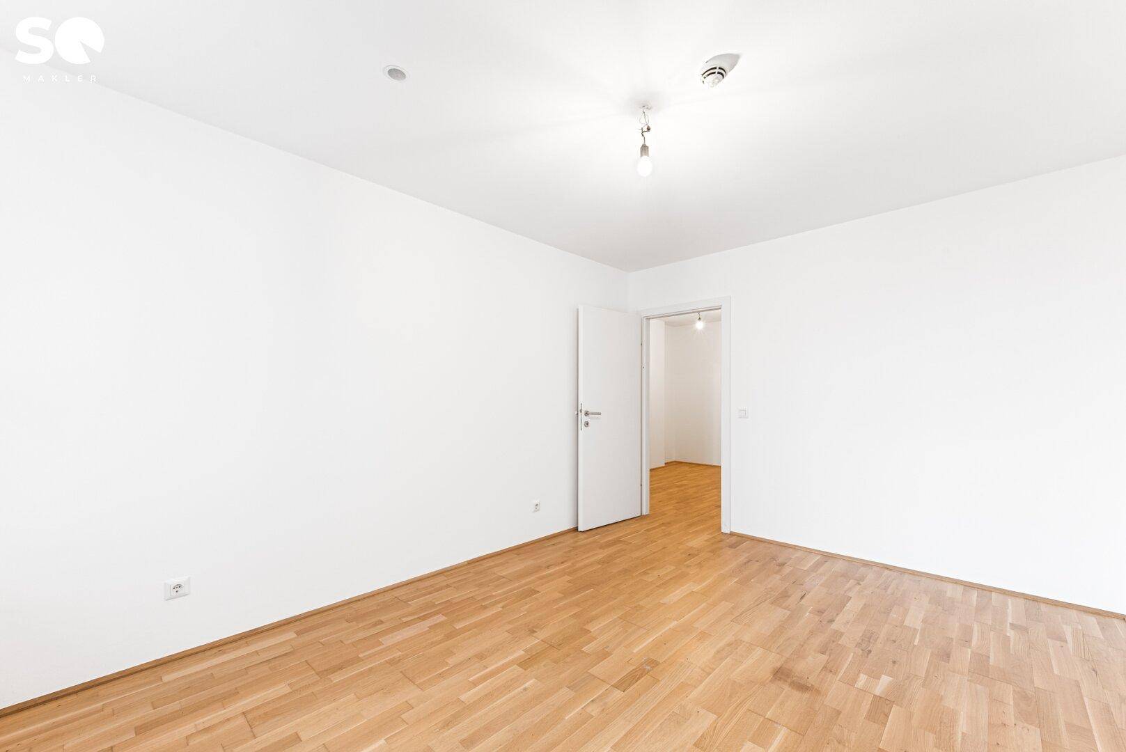 Heller, leerer Raum mit Parkettboden und weiß gestrichenen Wänden, mit Tür zu weiterem Zimmer.