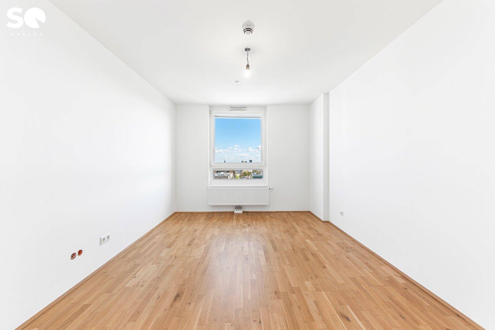 Leeres Schlafzimmer mit hellem Parkettboden und einem Fenster, das Tageslicht spendet.