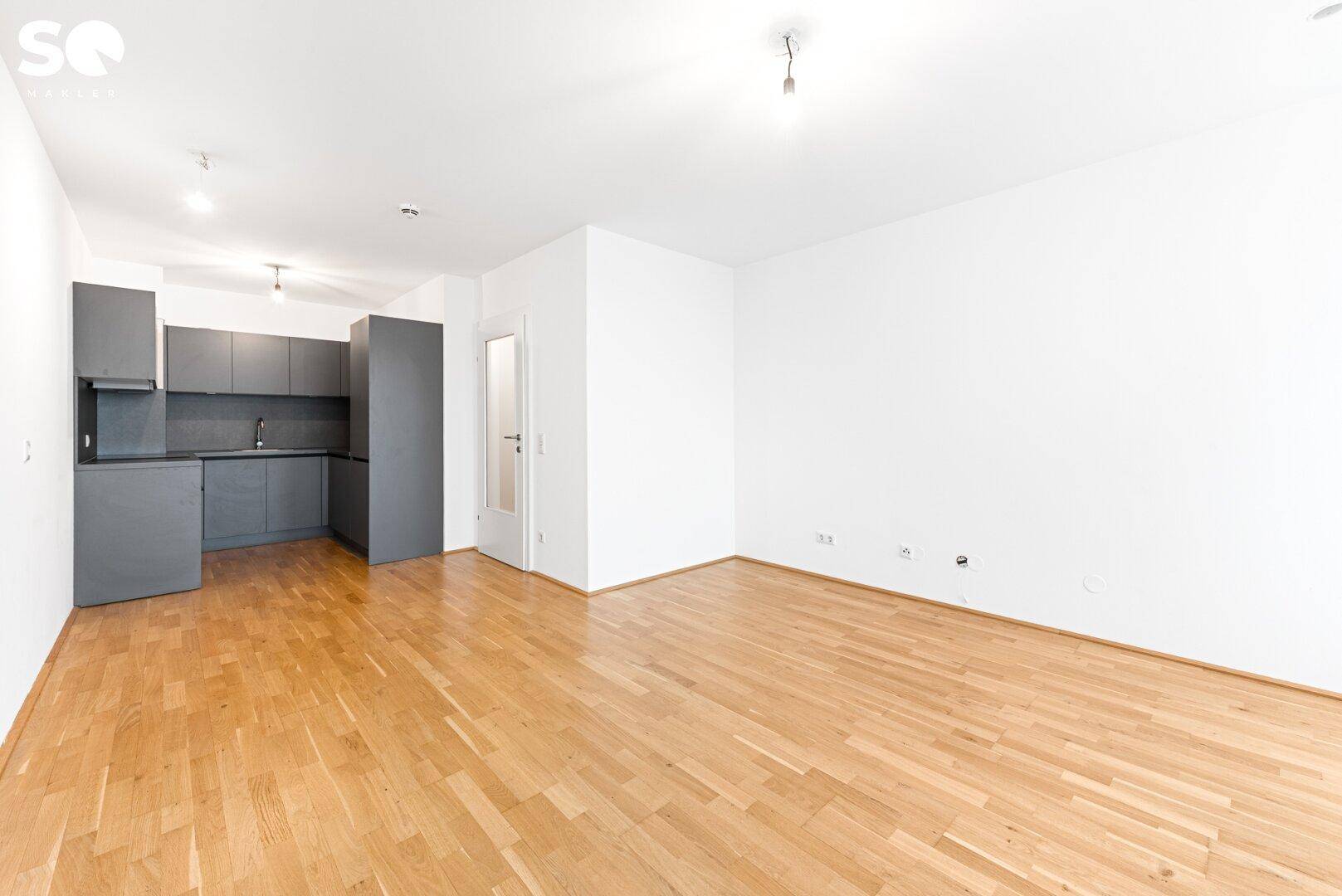 Geräumiger Wohnbereich mit Parkettboden und einer modernen, grauen Küchenzeile im Hintergrund.
