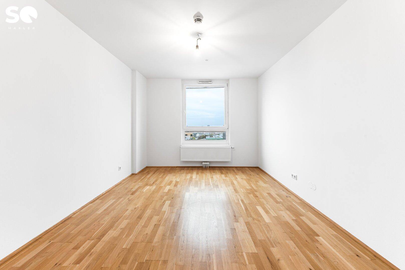 Leeres Zimmer mit Parkettboden, weißen Wänden und einem Fenster, das viel Tageslicht hereinlässt.