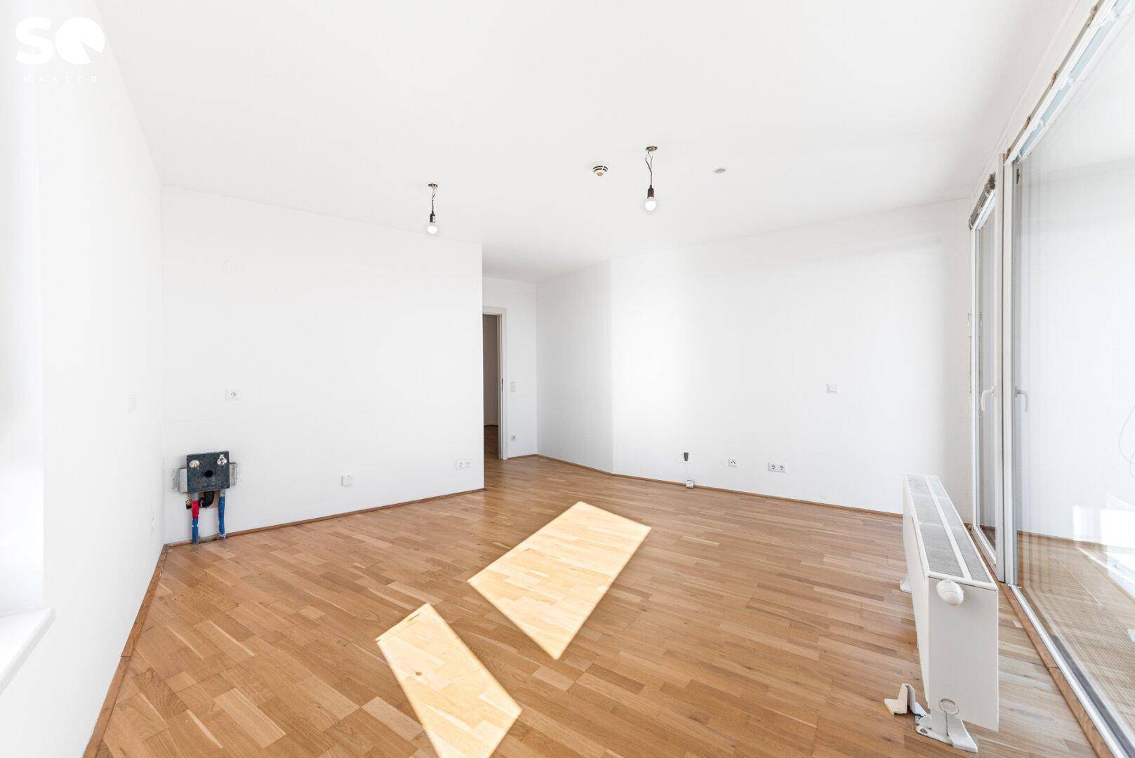 Heller Raum mit Parkettboden und sichtbaren Anschlüssen für eine zukünftige Kücheninstallation.