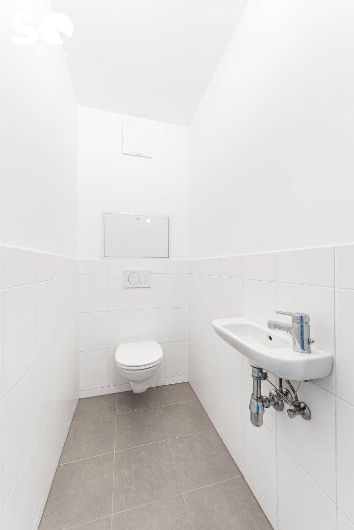 Separates WC mit Wand-WC, kleinem Waschbecken und weißen Fliesen, funktional und sauber.