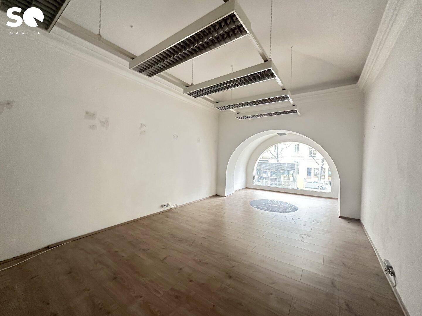 Geräumiger Raum mit Holzboden, Bogenfenster und Deckenleuchten, der viel natürliches Licht erhält.