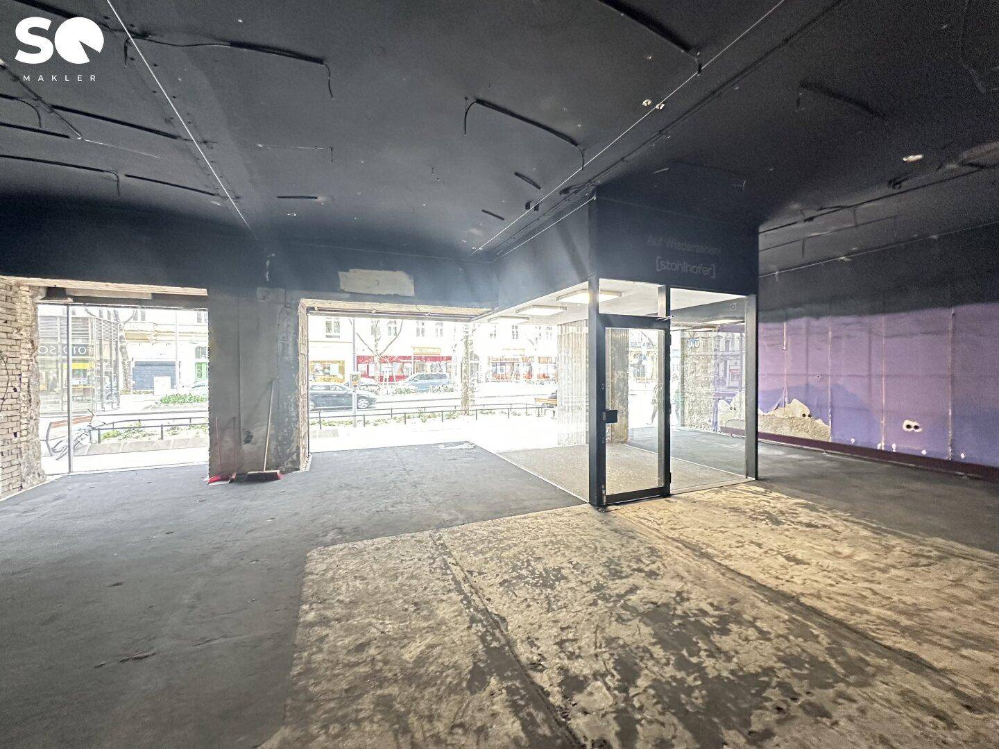 Geräumiger Geschäftsraum mit dunkler Decke und Boden, teilweise freigelegten Wänden und großer Fensterfront zur Straße.