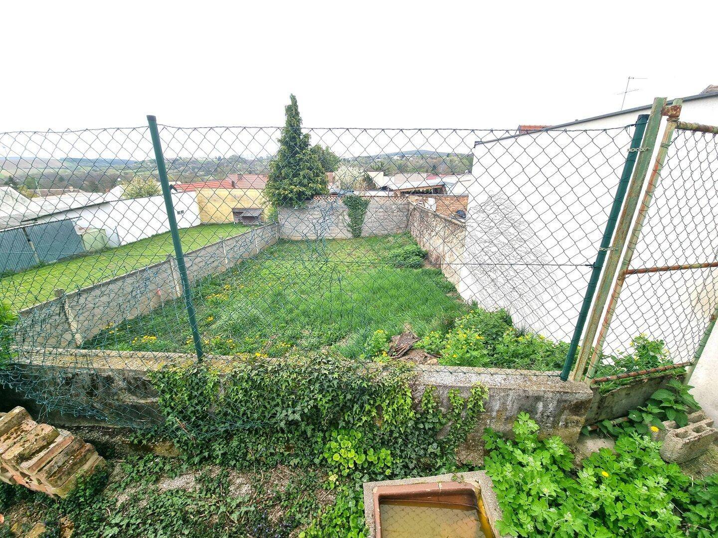 Eingezäunter Gartenbereich mit Rasen, Pflanzen und Blick auf die umliegende Landschaft und Häuser.