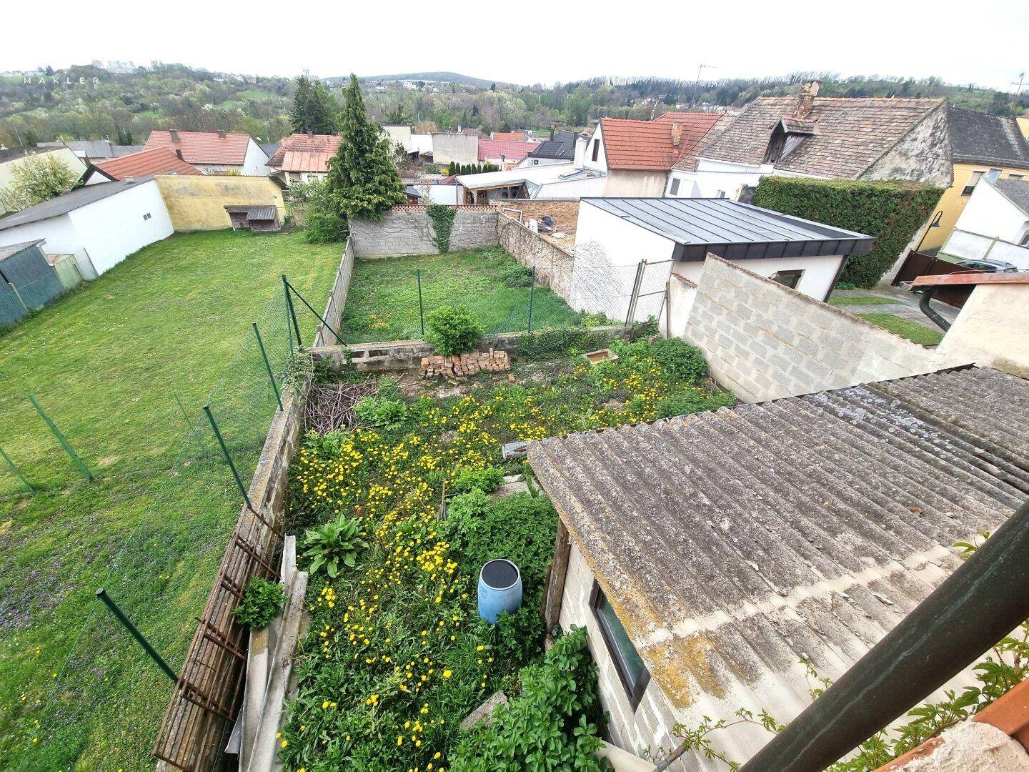 Vogelperspektive auf einen Gartenbereich mit Rasen, Blumen und umliegenden Häusern in der Ferne.