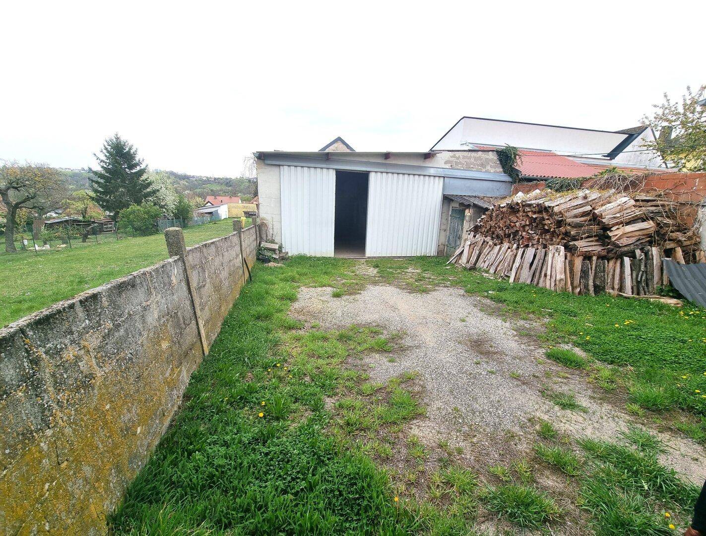 Außenbereich mit Grasfläche, Holzstapel und einer Scheune im Hintergrund, umgeben von Natur.