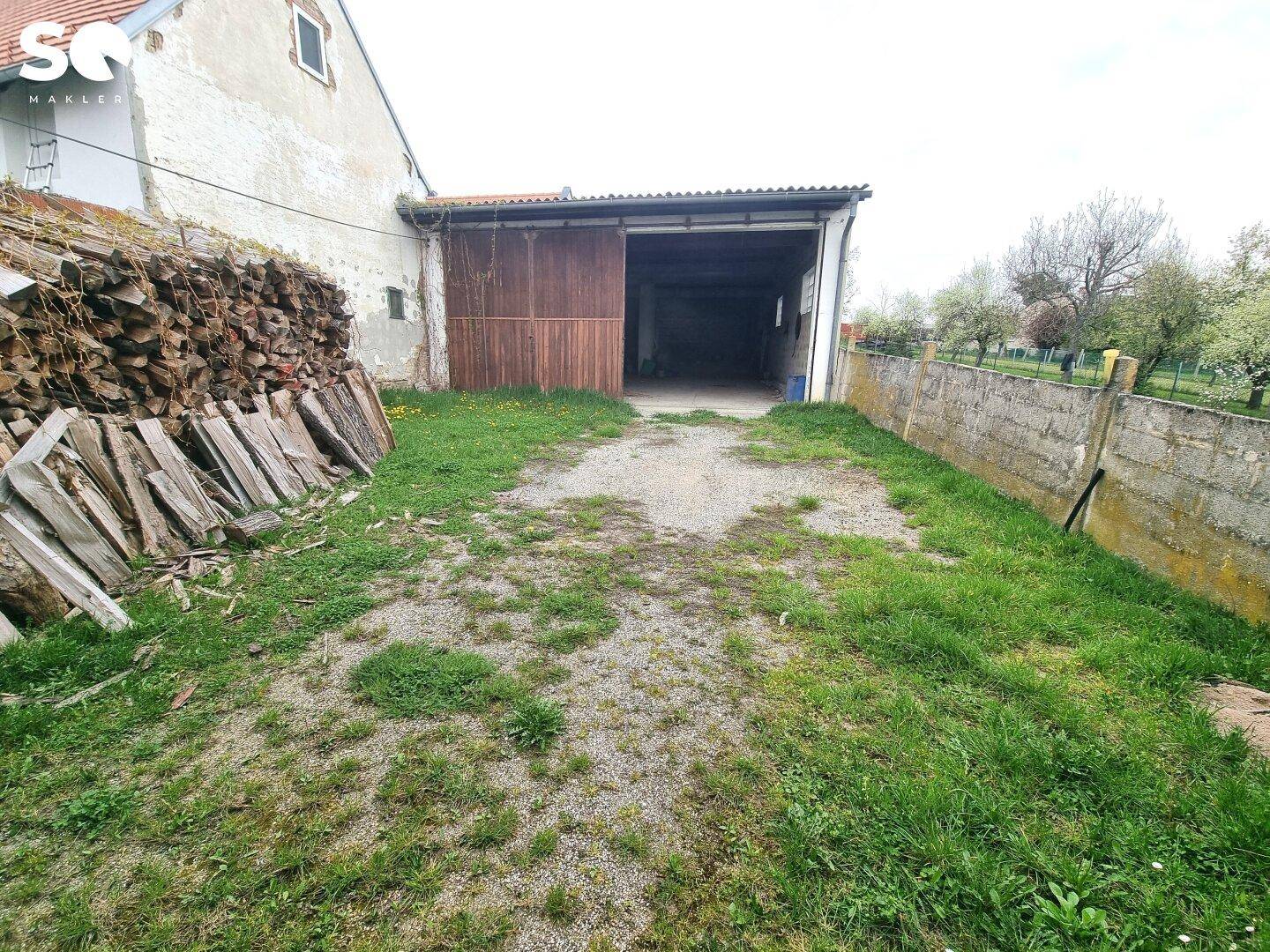 Außenbereich mit Grasfläche, Holzstapel und einer Scheune im Hintergrund, umgeben von Natur.