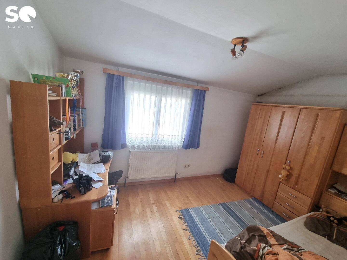 Schlafzimmer oder Arbeitszimmer mit Holzmöbeln, Parkettboden und einem Fenster mit blauen Vorhängen.