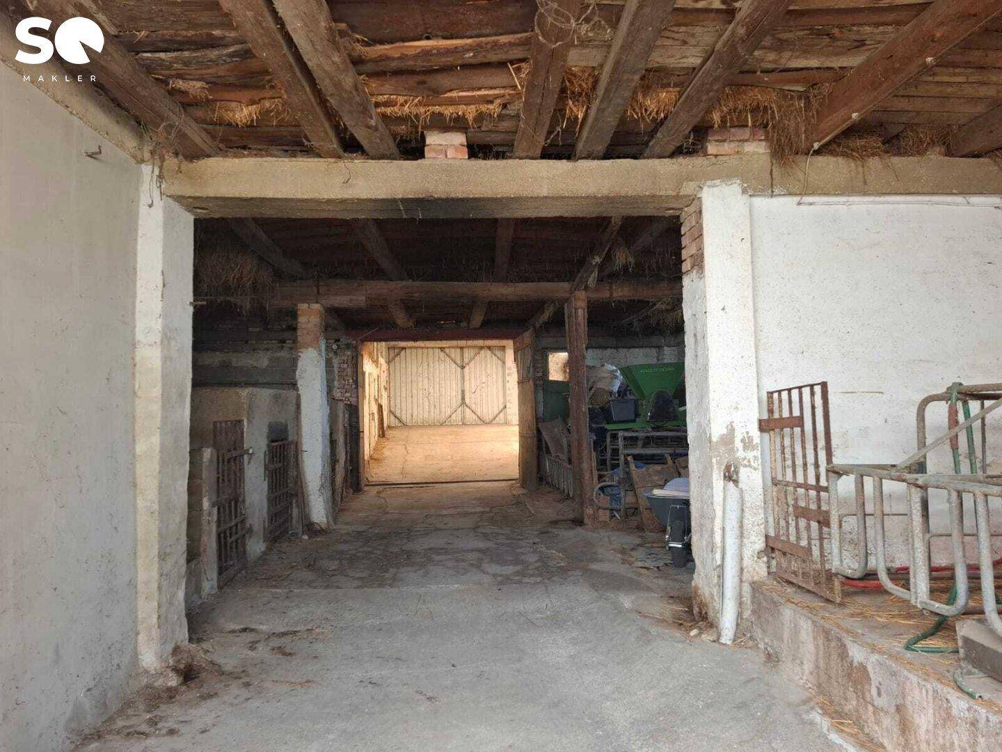 Innenansicht eines Stalls mit Holzbalken, Betonboden und Blick auf ein weiteres Tor im Hintergrund.