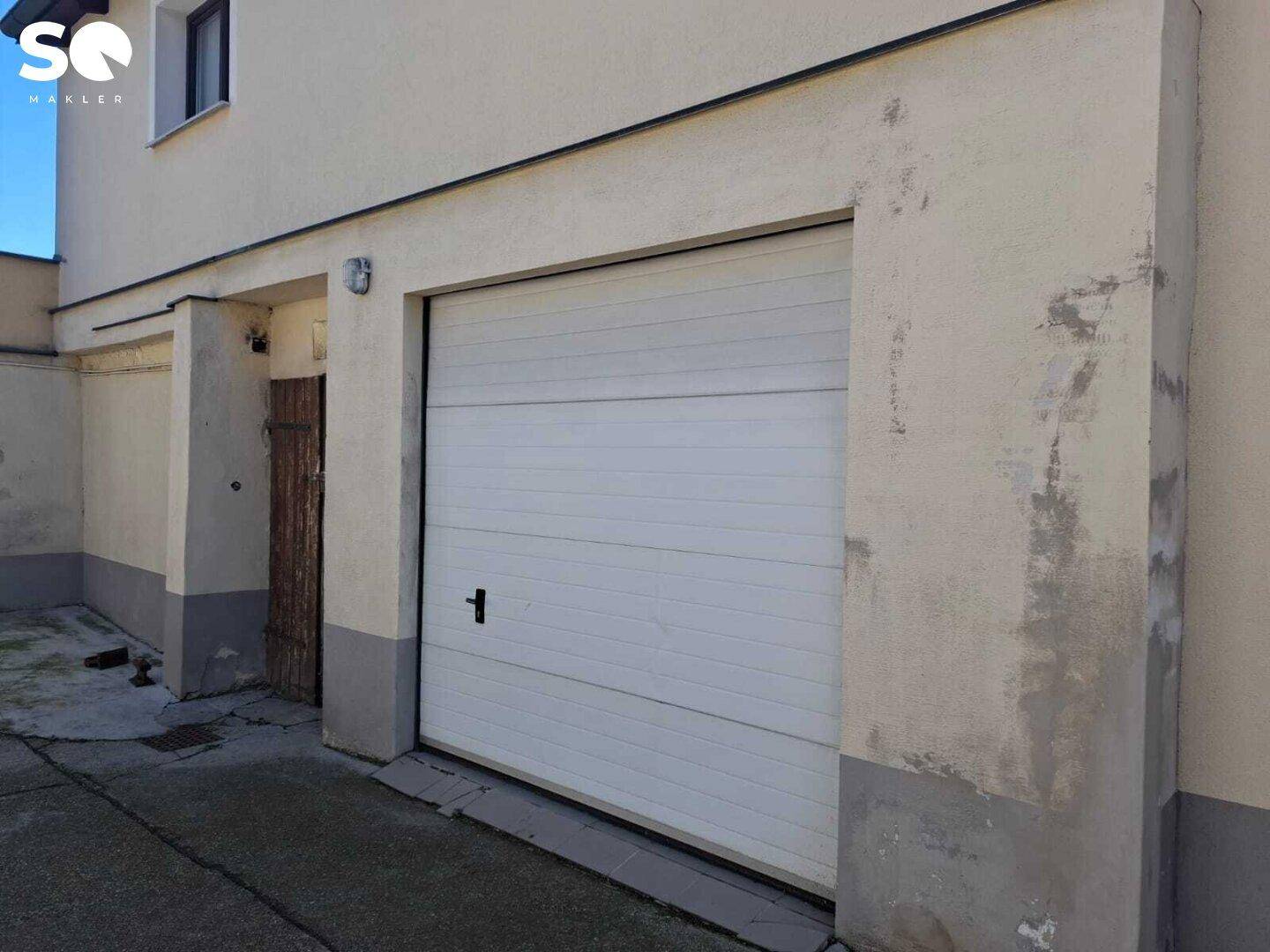 Garageneinfahrt mit einem weißen Rolltor und Putzfassade, die Renovierungsbedarf zeigt.