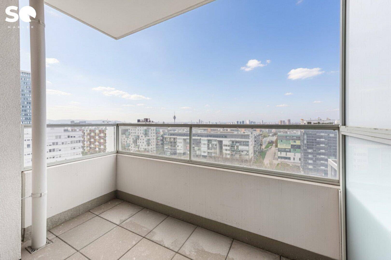 Balkon mit Glasbrüstung und weitem Blick über die Stadtlandschaft unter blauem Himmel.