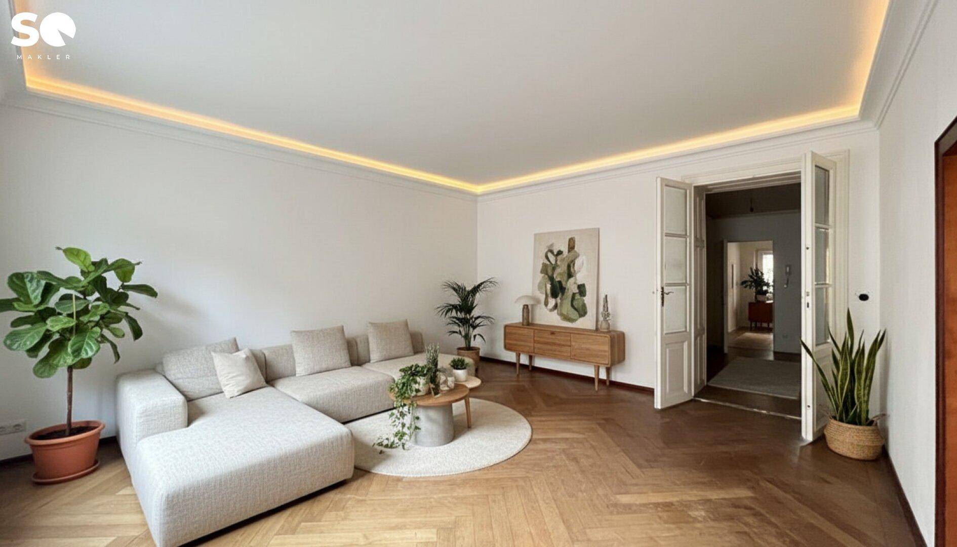 Modern eingerichtetes Wohnzimmer mit großem Sofa, Fischgrätparkett und indirekter Deckenbeleuchtung.