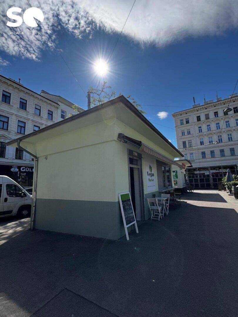 Vorderansicht des Gebäudes mit "Vegan Market" Schild und Außensitzgelegenheiten unter strahlendem Sonnenschein.