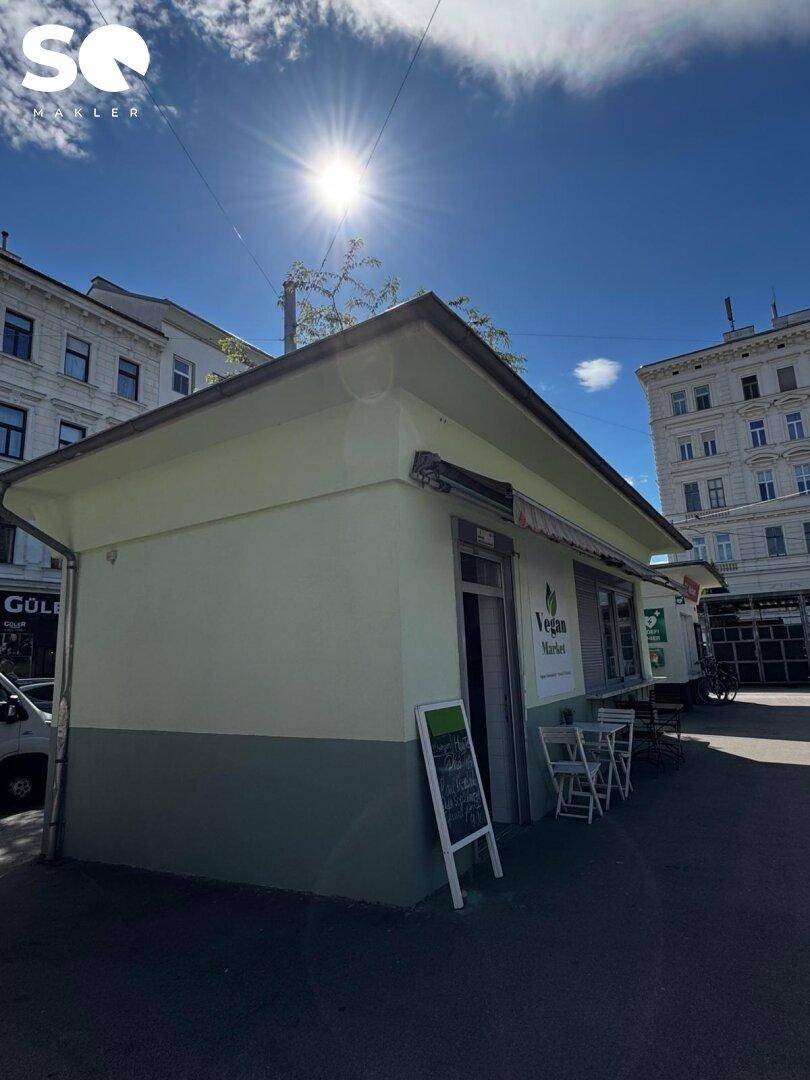 Das kleine Gebäude des "Vegan Market" mit Tischen im Freien, beleuchtet von der hellen Sonne am blauen Himmel.