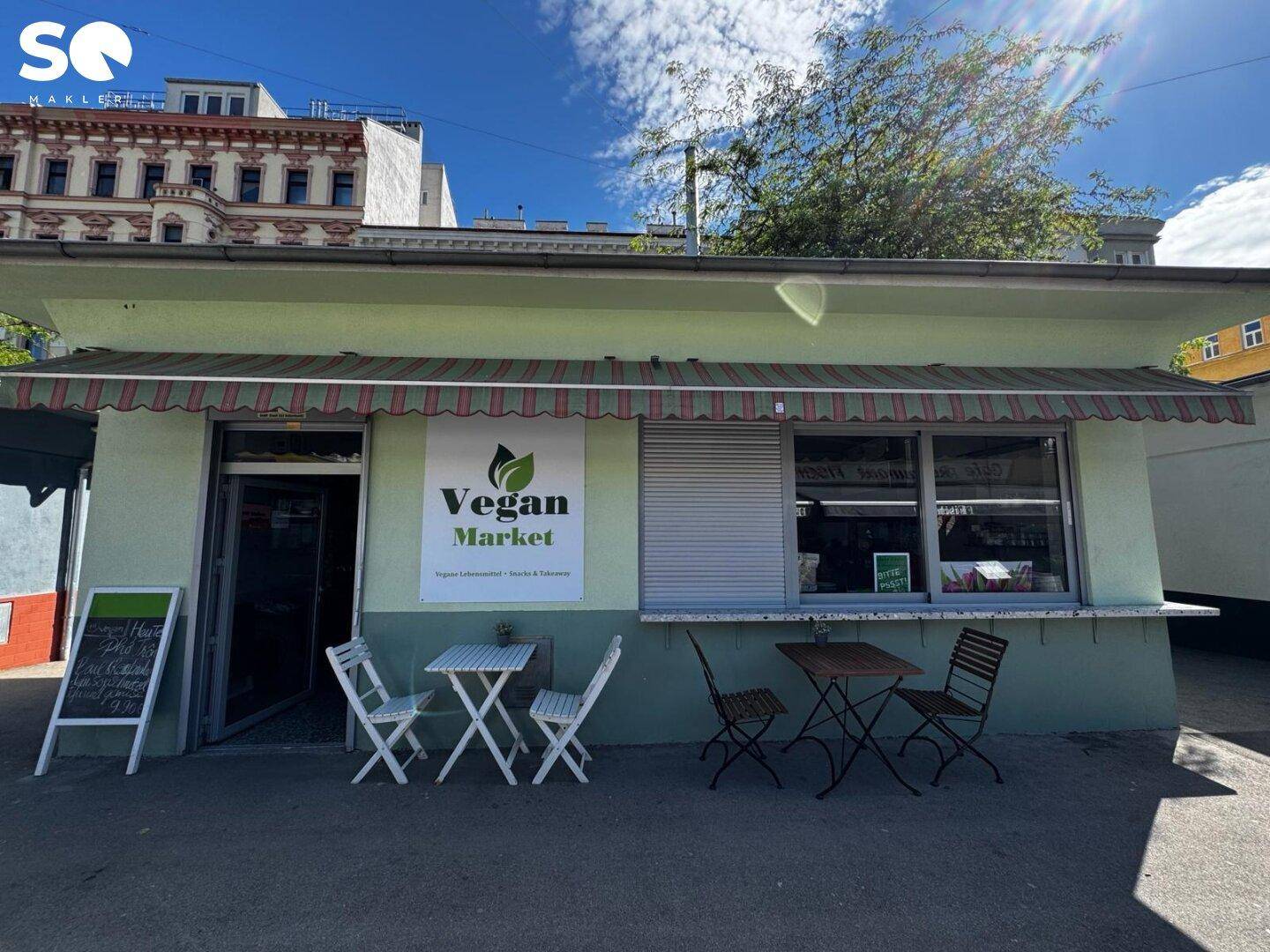 Direkte Frontansicht des "Vegan Market" mit geschlossener Jalousie und Tischen für Gäste im Außenbereich.