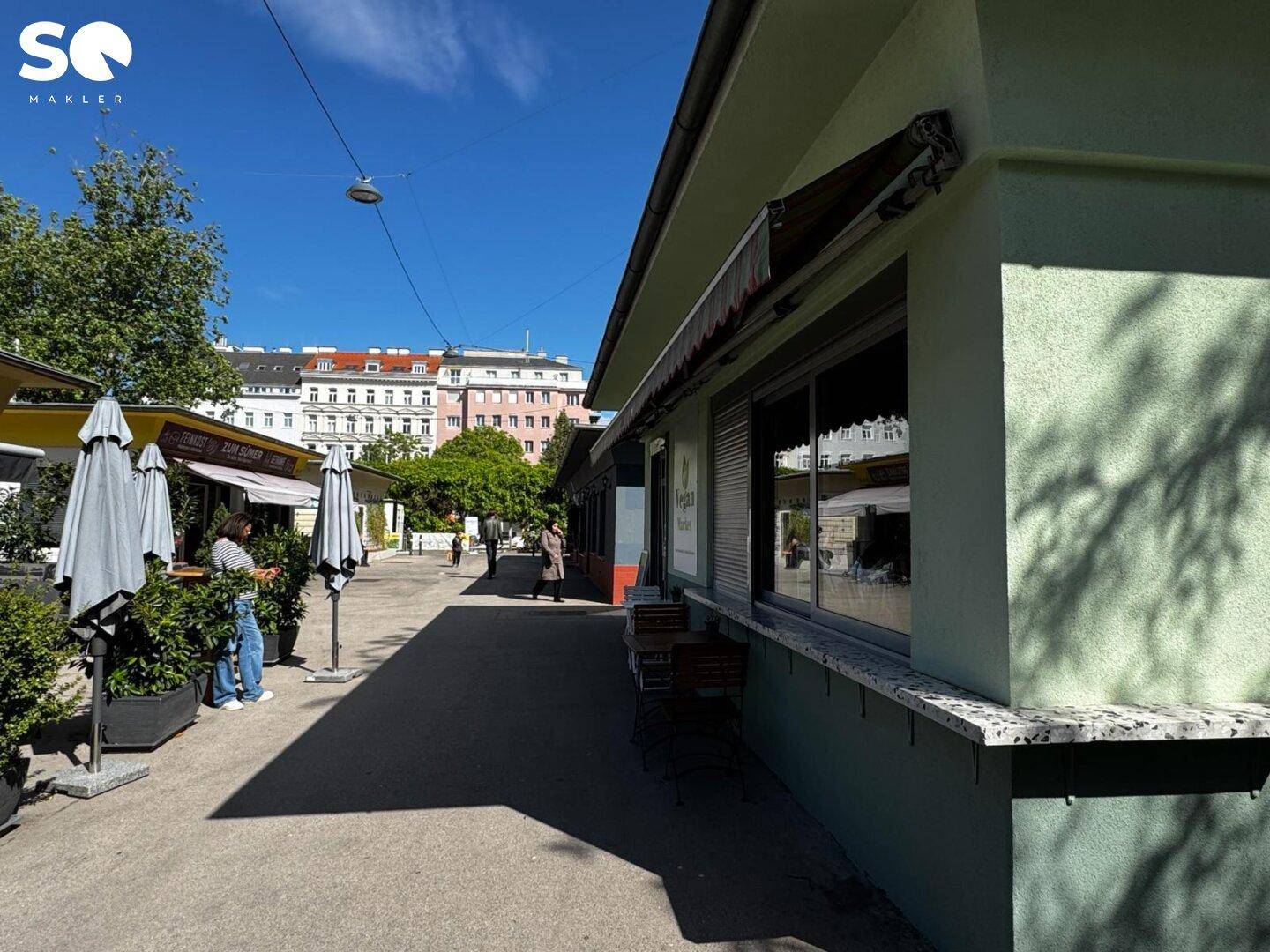 Seitenansicht des Gebäudes, blickt auf eine belebte Straße mit weiteren Marktständen und Cafés.