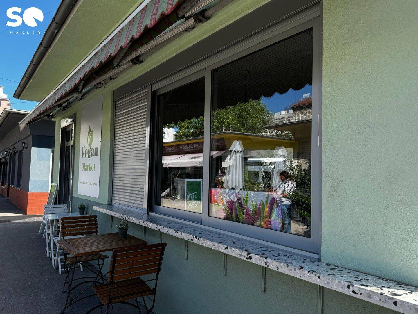 Nahaufnahme des Fensterbereichs mit Außentheke und Tischen, die die Umgebung im Glas widerspiegeln.